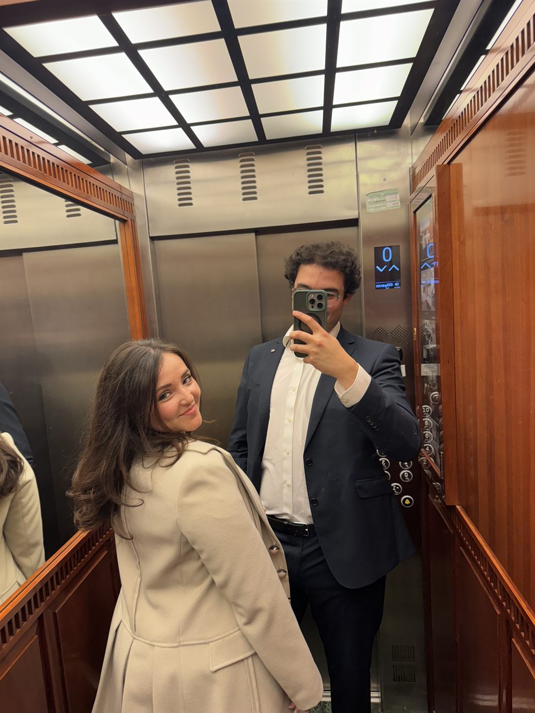 Selfie en el espejo de un ascensor, ella de abrigo claro y él de traje
