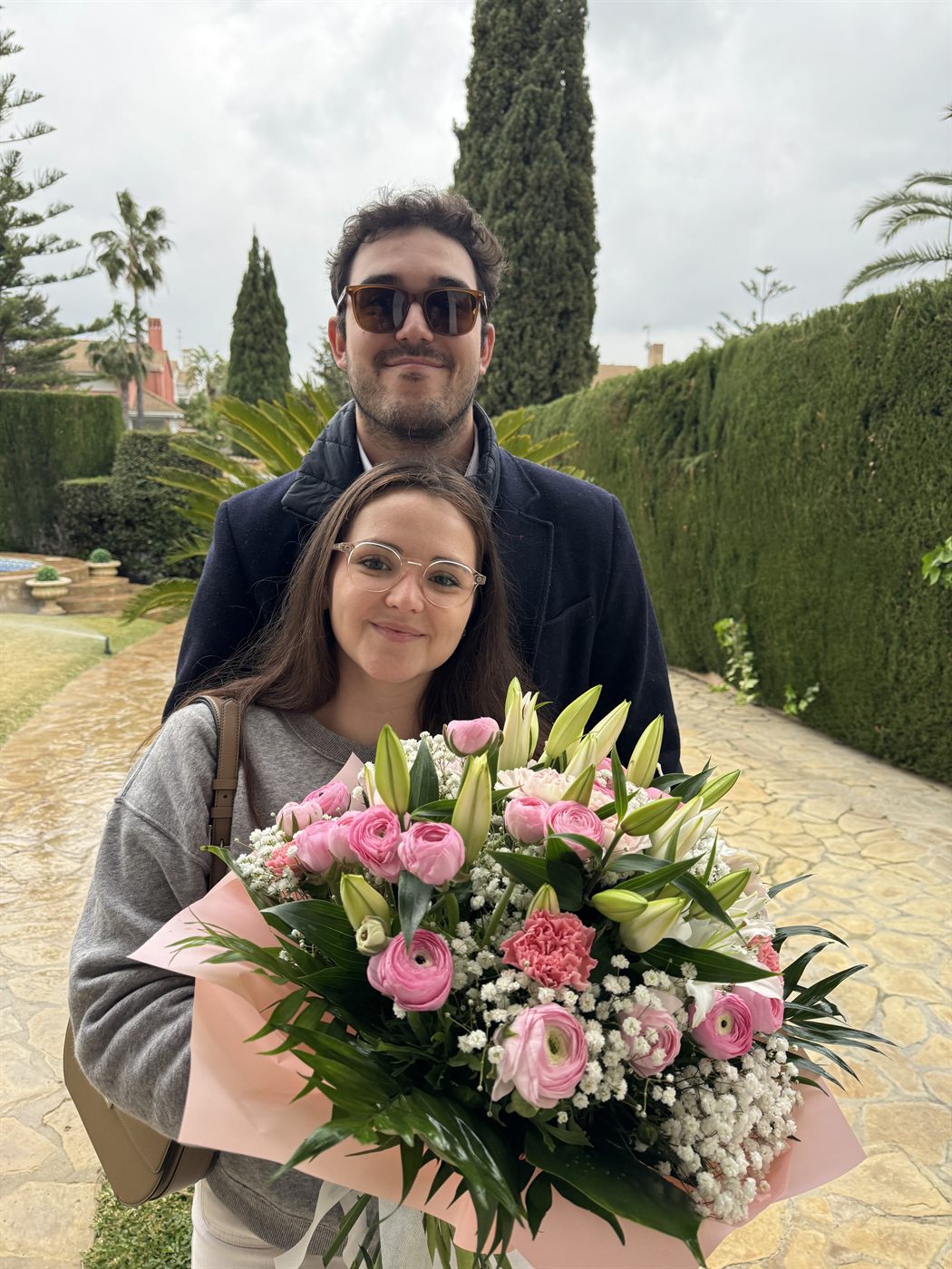 Los dos en un jardín, ella con un ramo grande de flores rosas y blancas